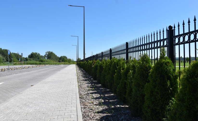 Metalowe ogrodzenie cmentarza z nasadzeniami zieleni, obok chodnik z kostki betonowej
