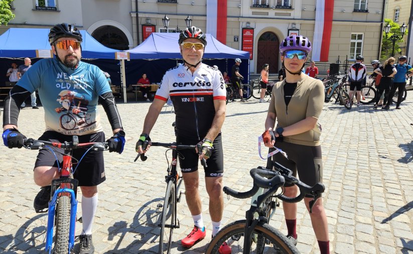 Trzech rowerzystów na rynku, za nimi niebieskie namioty