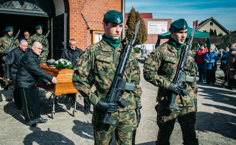 Na pierwszym planie żołnieże, na drugmi grupa osób wynosząca trumnę z kaplicy 