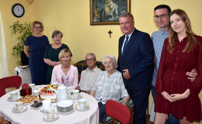 Stulatka siedząca przy stole, obok niej kilka uśmiechniętych osób 