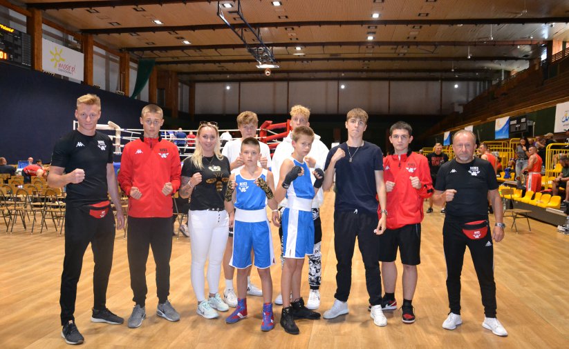 Grupa bokserów i bokserek w hali sportowej, po bokach trenerzy młodych zawodników