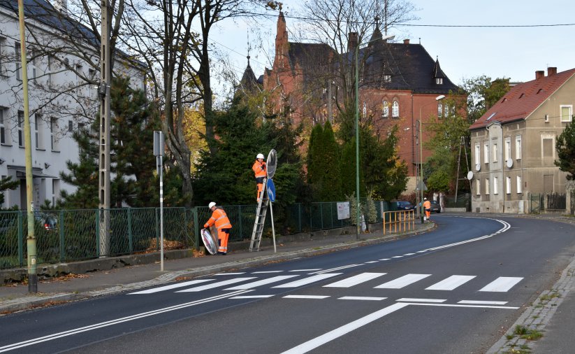 Droga, przejśćie dla pieszych, dwie osoby w pomarańczowych strojach montują znak drogowy