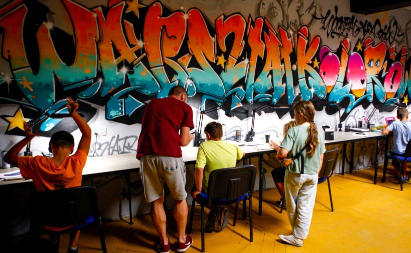 Na tle kolorowej ściany graffiti dzieci siedzą przy stołach i rysują, obok stoi instruktor