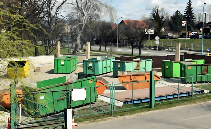 Duży plac przy drodze miejskiej, na placu poutawiane zielone kontenery na odpady