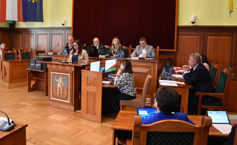 Sala obrad, za stopłem prezydialnym siedzą cztery sosoby, przed stołem mównica