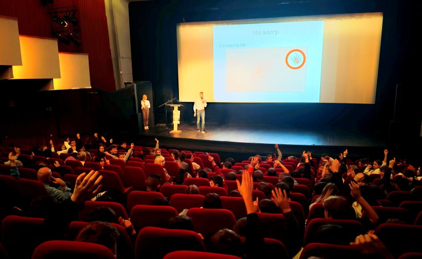 Sala Kinoteatru "Zbyszek", na scenie prowadzący wykład, nad fotelami kinowymi widać ręce zgłaszających się dzieci
