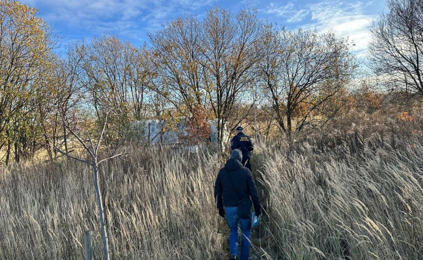 Teren nieużytków. Strażnik miejski i pracownik OPS-u zmierzają do przyczepy samochodowej ukrytej za drzewami