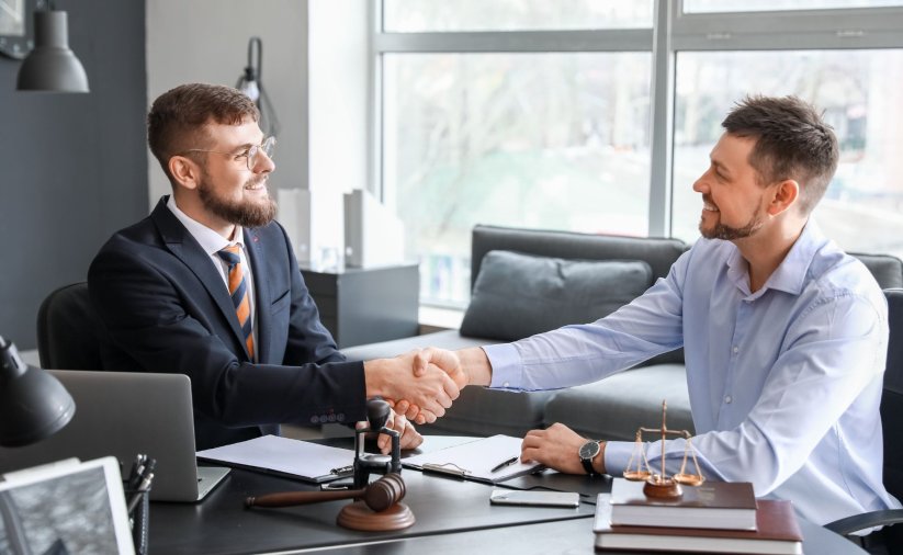 Mężczyzna w garniturze przy biurku, z dokumentami, tłumaczący coś drugiej osobie. Podają sobie wzajemnie dłoń.