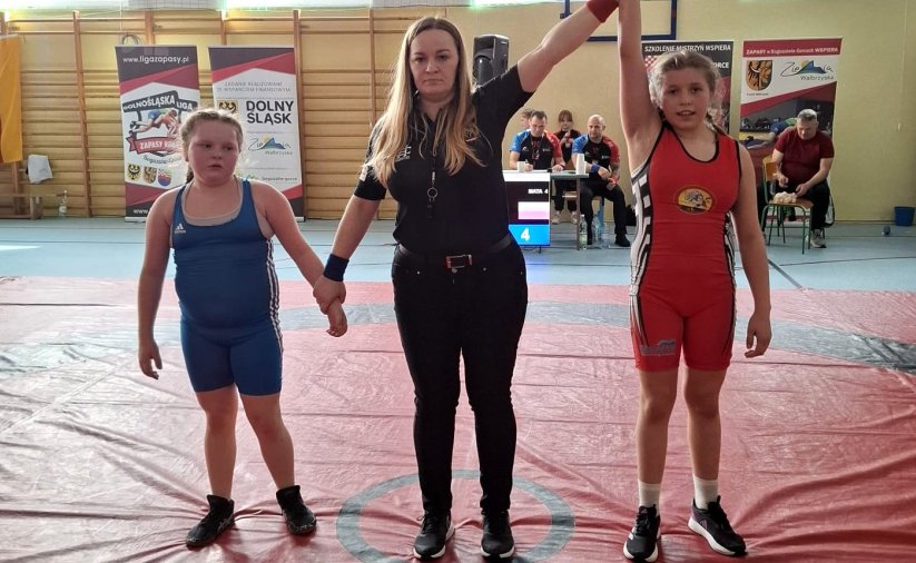 Dwie zawodniczki i sędzina na macie podczas ogłaszania wyniku