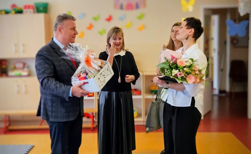 Burmistrz wręczający dyrektorce żłobka kwiaty i biały koszyk ze słodyczami