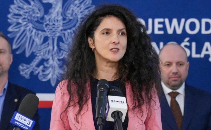 Wojewoda Dolnośląska przy mikrofonie, za nią dwóch mężczyzn na niebieskim tle