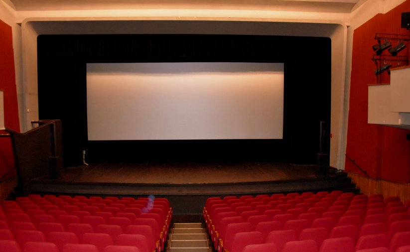 Biały ekran kinowy, przed ekranem scena i czerwone krzesełka, po bokach czerwone ściany