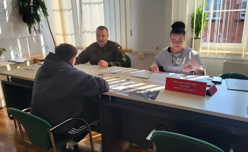 Żołnierz w mundurze i kobieta siedzący przy stole, przed nimi młody człowiek w bluzie z kapturem