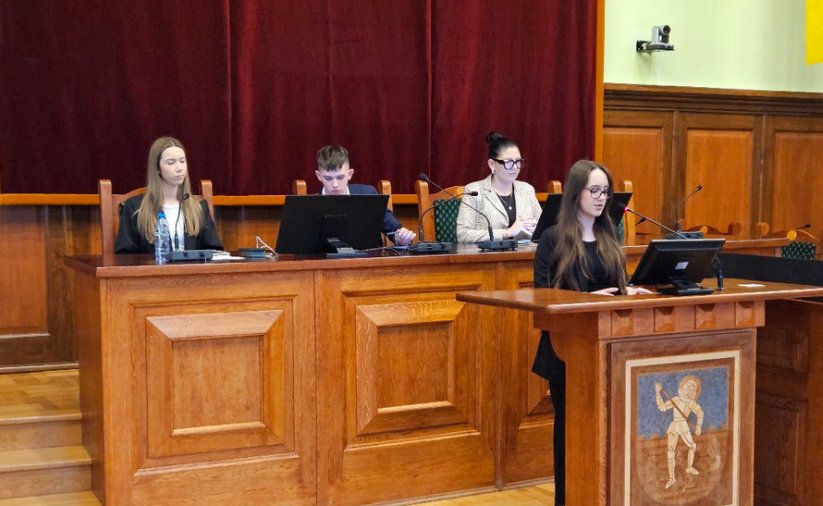Młodzieżowi radni, dziewczyna stojąca przy mównicy, chłopak i dwie dziewczyny siedzący za stołem prezydialnym