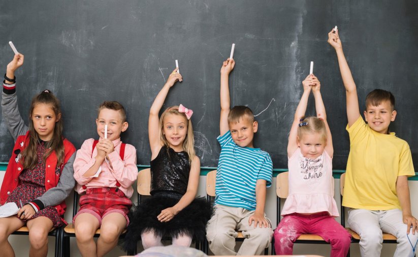 Trzech chłopców i trzy dziewczynki siedzą na krzesłach, za nimi tablica szkolna