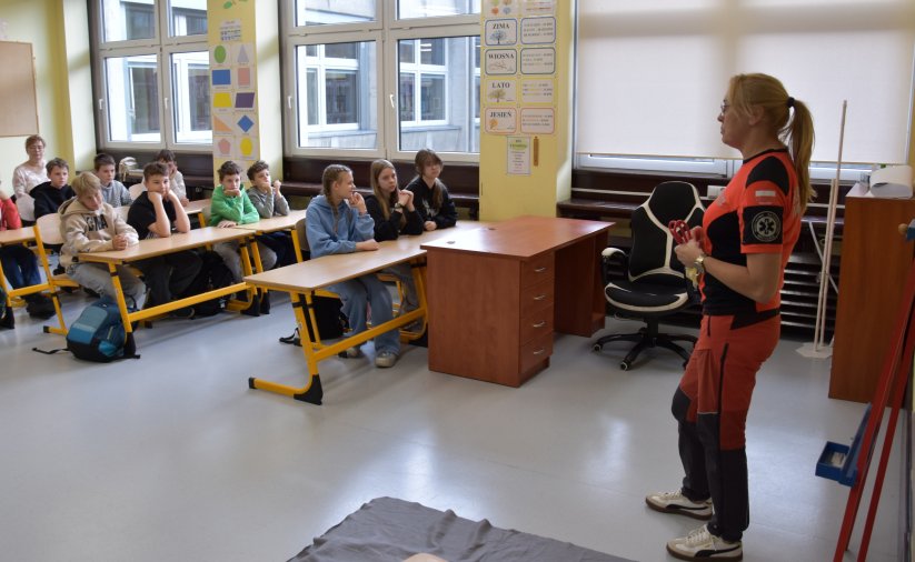 Pracownik oddziału ratunkowego w pomarańczowo-czarnym stroju stoi w klasie przed uczniami siedzącymi w ławkach