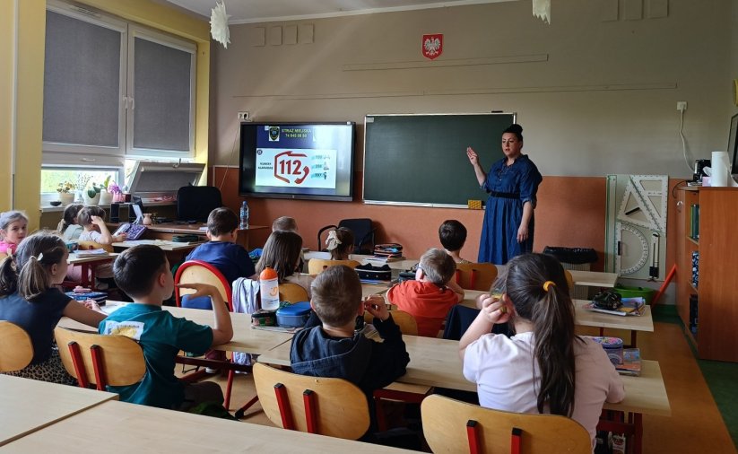 Klasa szkolna, młodzi uczniowie przy ławkach, przy tablicy pani w niebieskiej sukience