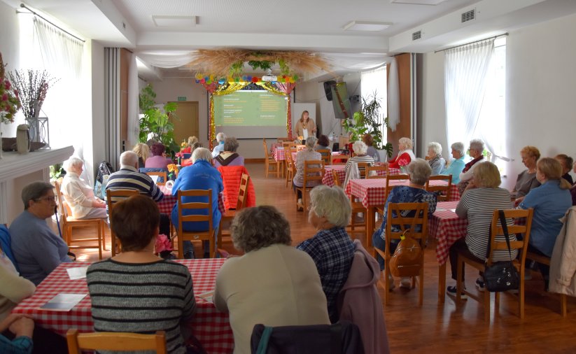 Duża sala, przy stołach siedzą seniorzy, na końcu sali kobieta przy dużym ekranie