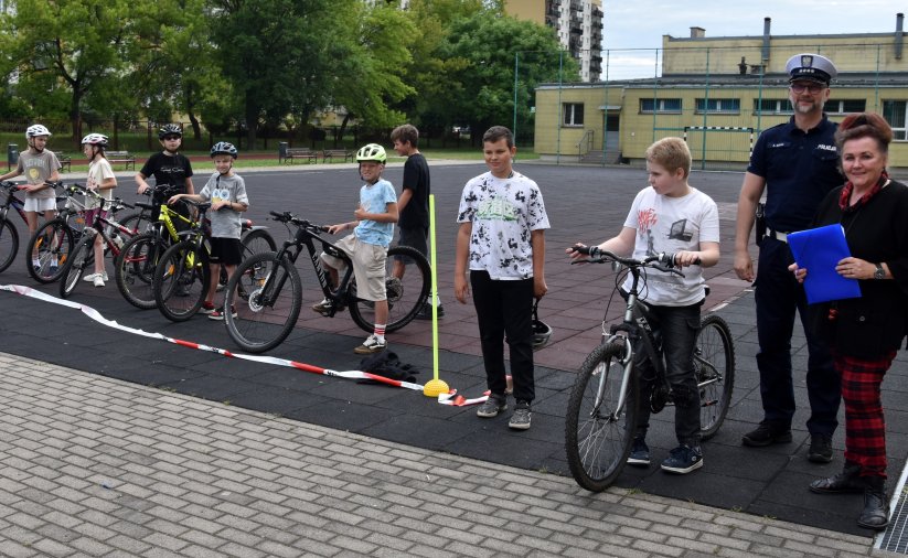 Grupa uczniów z rowerami ustawiona w szeregu, obok policjant i nauczycielka