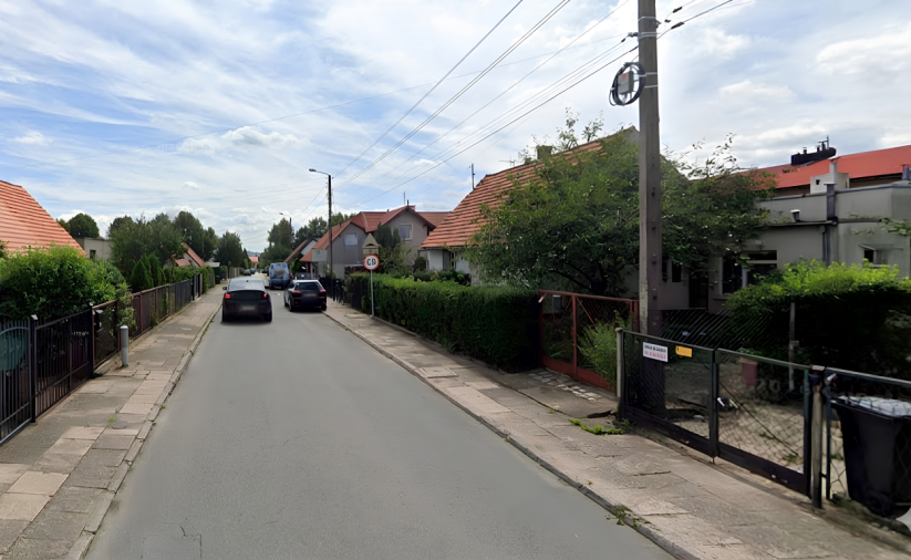 Uliczka, zaparkowane i jadące samochody, po bokach małe domki jednorodzinne