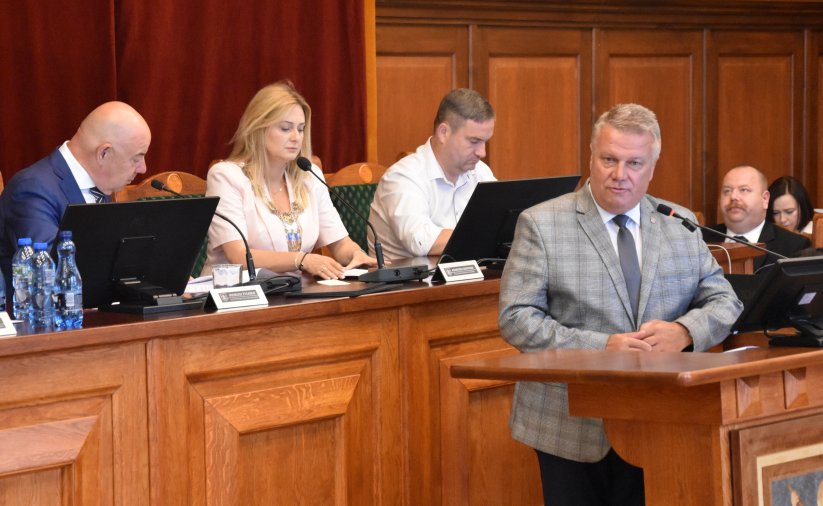 Burmistrz Dzierżoniowa przy mównicy, za nim przy stole prezydialnym przewodnicząca i dwóch wiceprzewodniczących rady