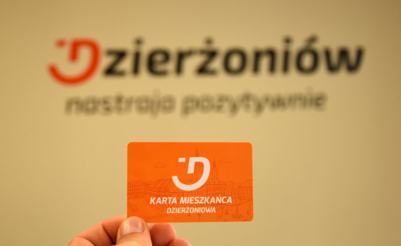 Dłoń trzymająca pomarańczową kartę mieszkańca na tle logotypu miasta