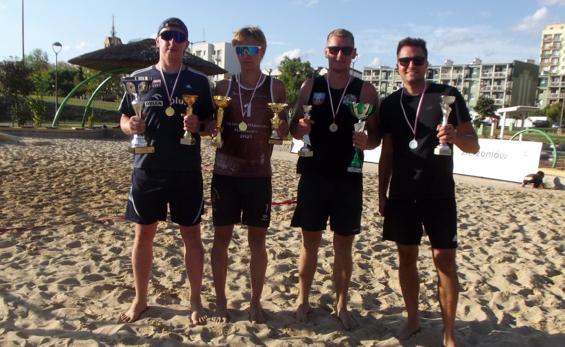 Czterech zawodników, każdy z pucharem i medalem, stojących na boisku do siatkówki plażowej, za nimi bloki osiedlowe