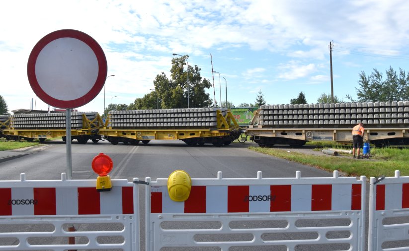 Przejazd kolejowy zagrodzony barierkami, na torach skład wiozący podkłady kolejowe