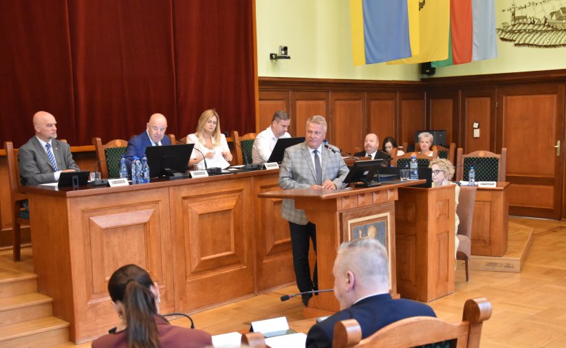 Sala obrad, przy mównicy burmistrz, za nim przewodnicząca i trójka wiceprzewodniczących przy stole prezydialnym