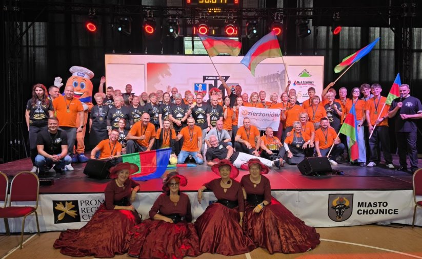duża grupa osób na scenia w czarnych i pomarańczowych koszulkach, na pierwszym planie cztery panie w bordowych sukniach i kapeluszacj