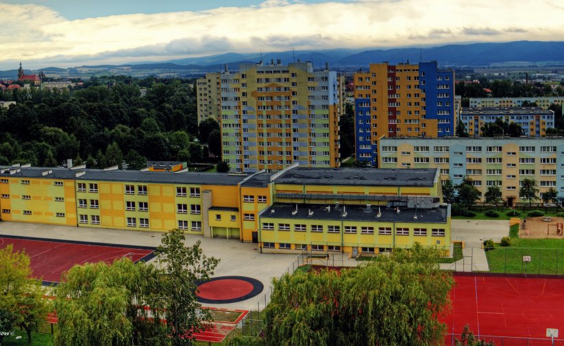 Trzypiętrowy budynek szkoły z żółtą elewacją, za nim wieżowce na tle gór