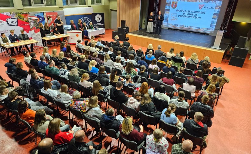 zdjęcie z góry sali wypełnionej osobami siedzącymi na krzesłach, na scenie dwie osoby, przed nimi slajd z rzutnika