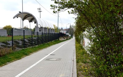 Chodnik i ścieżka rowerowa z kostki betonowej, po lewej za płotem zadaszenie trybun stadionu, po prawej zieleń