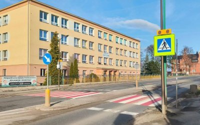 Szeroka droga, biało-czerwone przejście dla pieszych, za drogą duży trzypiętrowy budynek