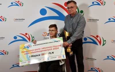 Młody chłopiec na wózku trzymający symboliczny czek, obok stoi dorosła osoba