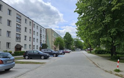 Droga osiedlowa z kostki betonowej, po lewej blok, po prawej duże drzewa