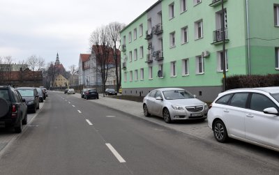 Droga asfaltowa, po bokach chodniki z zaparkowanymi autami, po prawej zielony trzypiętrowy blok
