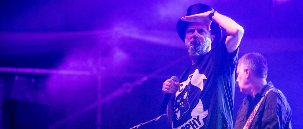 Wokalista zespołu na scenie w koszulce i cylindrze, za nim gitarzysta, niebieskie światło