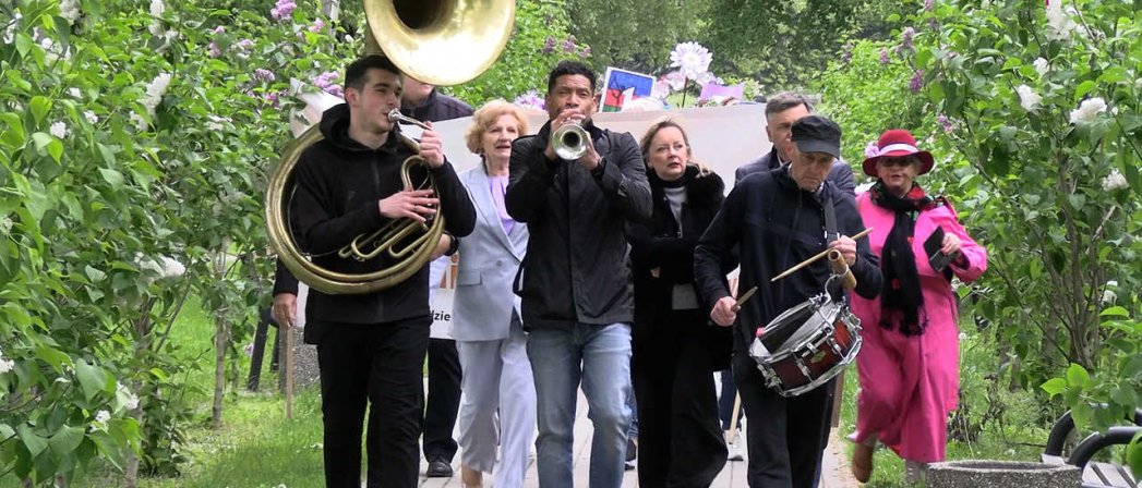 Aleją parkową idzie grając na instrumentach dwóch trębaczy i werblista, za nimi trzy kobiety, po bokach lilaki
