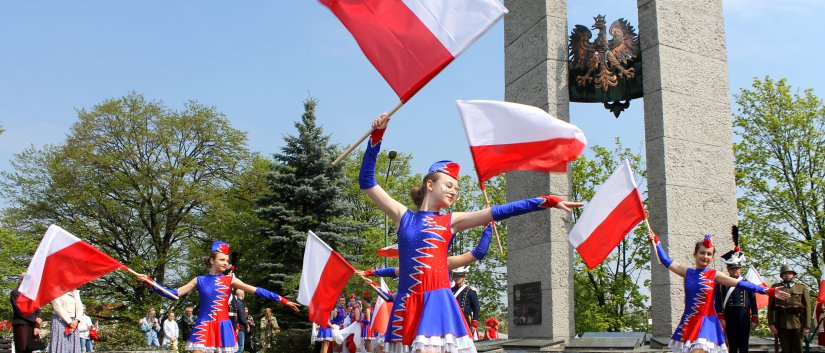 Dziewczyny z w niebieskich strojach z flagami Polski za nimi pomnik z godłem z metalu