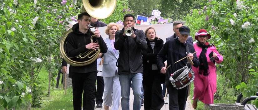 Aleją parkową idzie grając na instrumentach dwóch trębaczy i werblista, za nimi trzy kobiety, po bokach lilaki