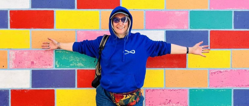 Uśmiechnięta Roksana Jędrzejewską-Wróbel w niebieskiej bluzie z kapturem, z rozłożonymi na boki rękoma na tle ściany z kolorowych poziomych prostokątów